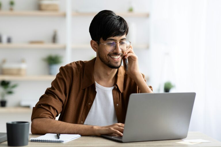 Cheerful indian guy businessman with laptop having phone conversation